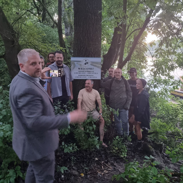 Jedlička takes a picture of the BORDER CROSSING LIBERLAND-CROATIA sign. Houseboat visible on the right